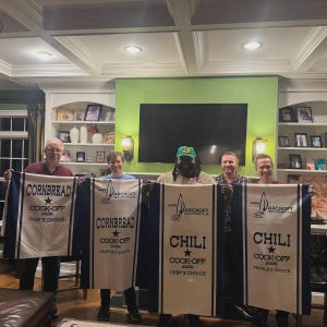 Puede incluir: Un grupo de personas sostienen pancartas para un concurso de cocina. Las pancartas blancas con bordes azules incluyen las palabras "CORNBREAD" y "CHILI", adem&aacute;s de "COOK-OFF 2026" y "CHEF'S CHOICE" o "PEOPLE'S CHOICE".