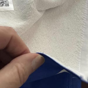 May include: Close-up of a white terry cloth towel with a blue border. The towel is folded, revealing the blue edge and a portion of the white terry cloth. A hand is partially visible.