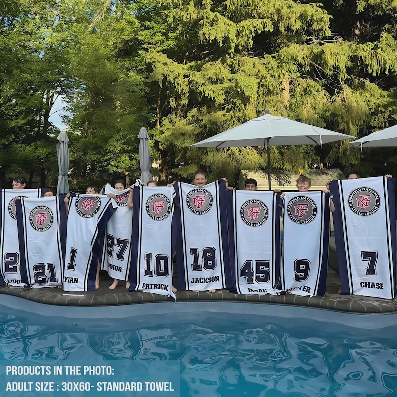 Puede incluir: Un grupo de personas sostienen toallas blancas y azul marino con el logotipo de Fairfield National Little League. Cada toalla tiene un n&uacute;mero y un nombre diferentes. Las toallas miden 76 x 152 cm.
