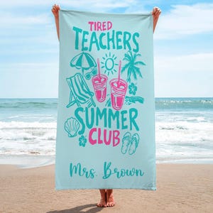 May include: A light blue beach towel with the words "Tired Teachers Summer Club" in teal and pink. The towel features illustrations of a beach umbrella, sunglasses, drinks, a chair, and a seashell. The name "Mrs. Brown" is printed at the bottom.