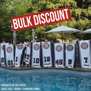 Puede incluir: Un grupo de personas sostienen toallas est&aacute;ndar blancas y azul marino con el texto "BULK DISCOUNT" en una pancarta roja. Cada toalla tiene un logotipo y un n&uacute;mero con un nombre. Las toallas miden 76x152 cm.