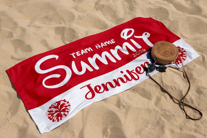May include: A red and white beach towel with the name "Summit" in large white script, the name "Jennifer" in red script, and a pom-pom graphic. A woven bag and sunglasses rest on the sand next to the towel.