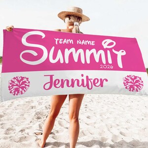 May include: A pink and white personalized beach towel with the name "Summit" in large white script, along with "Jennifer" and the year "2020". The towel features pom-pom graphics on each end.