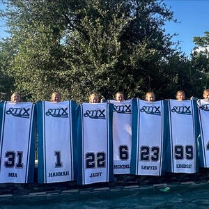 Puede incluir: Un grupo de chicas sosteniendo camisetas blancas y turquesas con n&uacute;meros y nombres. Las camisetas tienen la palabra "STIX" en la parte superior. Los n&uacute;meros incluyen 00, 12, 31, 1, 22, 6, 23, 99, 42, 15 y 24. Los nombres est&aacute;n impresos debajo.