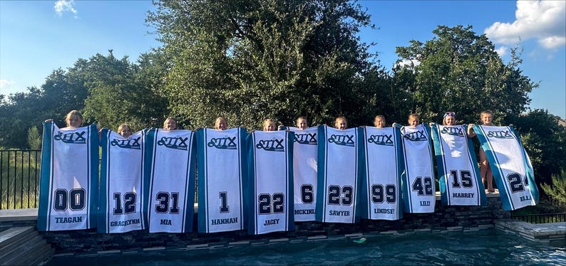 Puede incluir: Un grupo de chicas sosteniendo camisetas blancas y turquesas con n&uacute;meros y nombres. Las camisetas tienen la palabra "STIX" en la parte superior. Los n&uacute;meros incluyen 00, 12, 31, 1, 22, 6, 23, 99, 42, 15 y 24. Los nombres est&aacute;n impresos debajo.