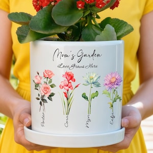 Op de afbeelding: Een witte keramische Mom's Garden plantenbak met bijpassende schotel, gevuld met een rood bloeiende plant. De plantenbak heeft bloemenillustraties en de tekst "Mom's Garden Love Grows Here".