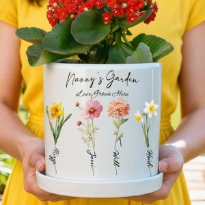 May include: A white ceramic flower pot with the text "Nanny's Garden Love Grows Here" and floral illustrations. The pot holds a plant with red flowers and green leaves. The pot is held by a person wearing a yellow dress.