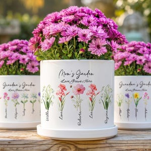 Może przedstawiać: Trzy białe ceramiczne donice z żywymi różowymi kwiatami. Donice są ozdobione ilustracjami kwiatowymi oraz napisami "Mom's Garden" i "Grandma's Garden". Każda donica ma pasujący spodek.