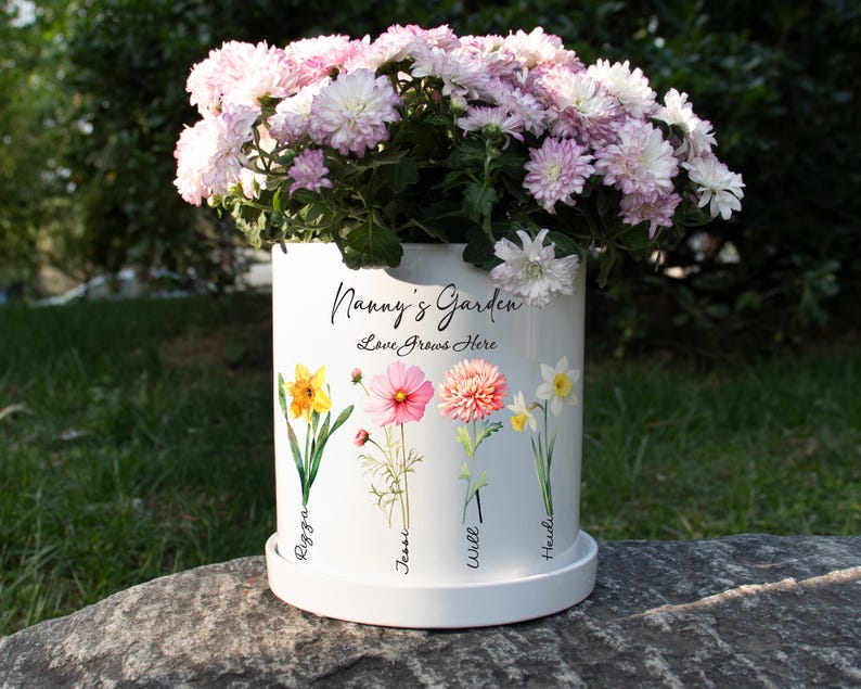 May include: A white ceramic flower pot with the text "Nanny's Garden Love Grows Here" and floral illustrations. The pot is filled with pink and white chrysanthemums. The names "Regga", "Jessi", "Will", and "Heidi" are printed below the flowers.