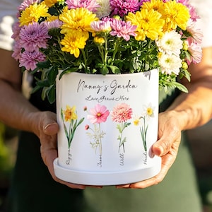 May include: A white ceramic flower pot filled with colorful chrysanthemums. The pot is decorated with floral illustrations and the text "Nanny's Garden Love Grows Here". The pot is held by a person.
