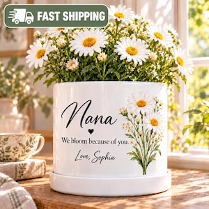 May include: A white ceramic cylindrical planter with a matching saucer, featuring a bouquet of white daisies and the black text "Nana We bloom because of you. Love, Sophia" with a small heart icon printed on the front surface.