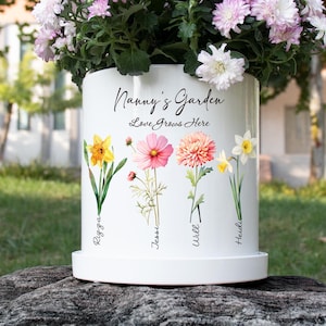 May include: White ceramic flower pot with the text "Nanny's Garden Love Grows Here" and illustrations of colorful flowers. The pot has a matching saucer and is filled with pink and white flowers.