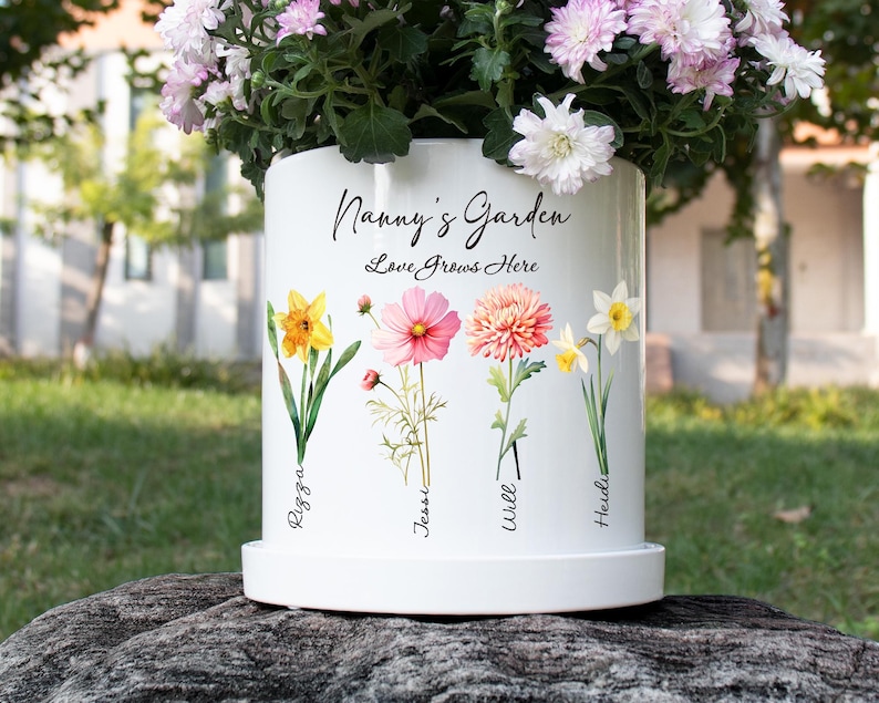 May include: White ceramic flower pot with the text "Nanny's Garden Love Grows Here" and illustrations of colorful flowers. The pot has a matching saucer and is filled with pink and white flowers.