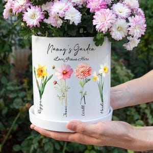 May include: White ceramic flower pot with the text "Nanny's Garden Love Grows Here". The pot features floral illustrations and names. The pot is filled with pink and white flowers.