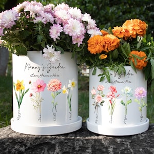 May include: Two white ceramic flower pots with floral designs and the text "Nanny's Garden" and "Mom's Garden". The pots are filled with colorful flowers, including pink, orange, and yellow blooms. The text "Love Grows Here" is also visible.