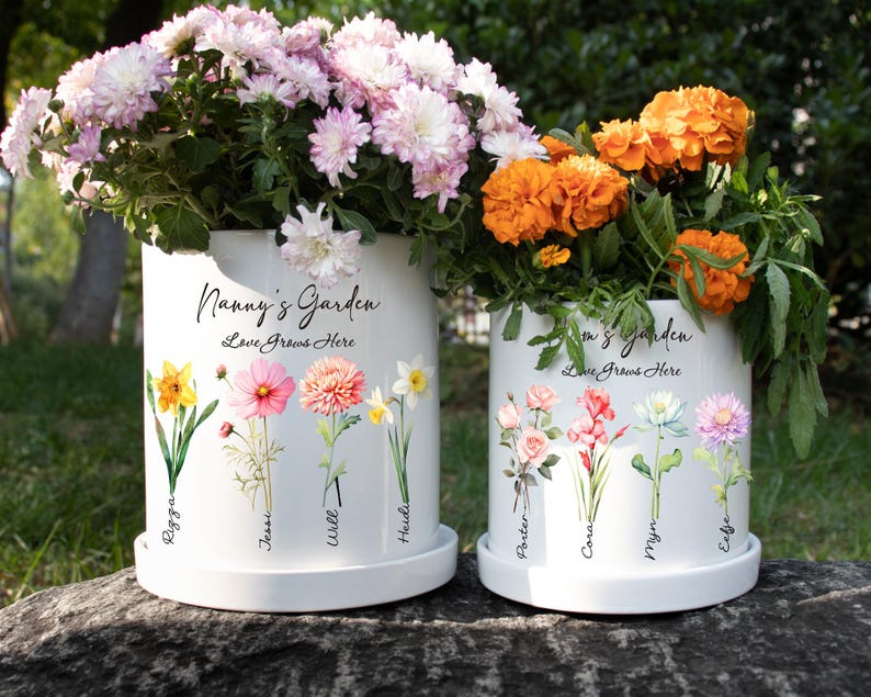 May include: Two white ceramic flower pots with floral designs and the text "Nanny's Garden" and "Mom's Garden". The pots are filled with colorful flowers, including pink, orange, and yellow blooms. The text "Love Grows Here" is also visible.