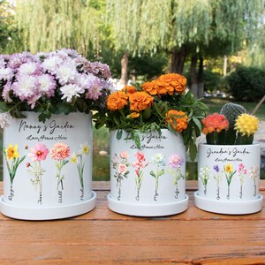 May include: Three white ceramic flower pots with floral designs and the text "Nanny's Garden" and "Grandma's Garden". The pots are filled with colorful flowers, including purple, orange, and yellow blooms. The pots sit on a wooden surface.