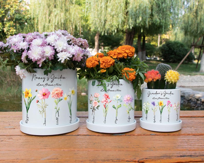 May include: Three white ceramic flower pots with floral designs and the text "Nanny's Garden" and "Grandma's Garden". The pots are filled with colorful flowers, including purple, orange, and yellow blooms. The pots sit on a wooden surface.