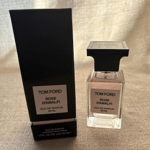 May include: A perfume bottle and box set. The perfume bottle is square with a black cap and a light pink liquid. The box is black with white text that reads "TOM FORD ROSE D'AMALFI EAU DE PARFUM 50 ML". The bottle is 1.7 fl oz.