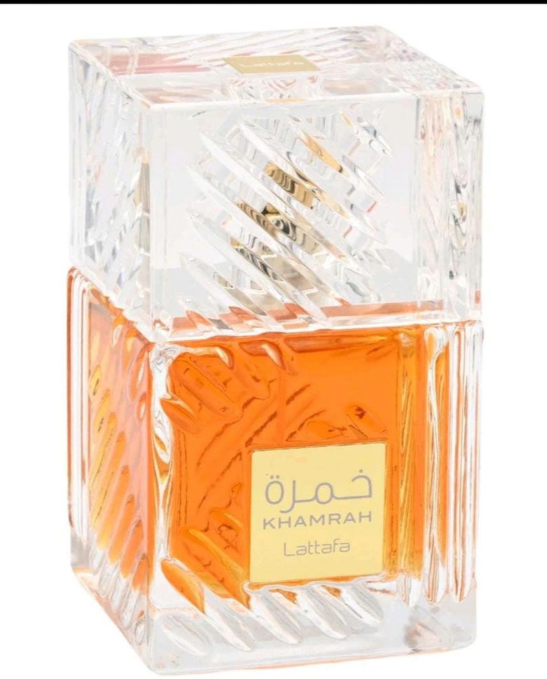 May include: A square glass perfume bottle with a clear, textured, faceted exterior and a matching heavy cap. The bottle contains an amber-colored liquid and features a gold-colored square label on the front with the text Khamrah Lattafa.
