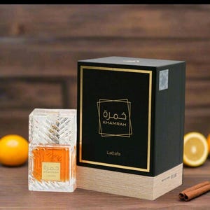 May include: A rectangular glass perfume bottle with amber liquid and a textured surface next to a black and tan box. The box features gold trim, the text Khamrah in English and Arabic, and the brand name Lattafa. The bottle label also displays the text.