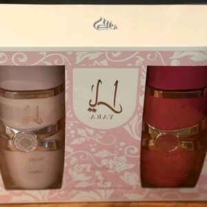 Peut inclure: Coffret cadeau avec deux contenants cylindriques. L'un est rose clair, l'autre rouge. Les deux ont des accents dor&eacute;s et une bande d&eacute;corative. La bo&icirc;te est blanche avec des motifs floraux roses et le mot "Yara" en &eacute;criture arabe.