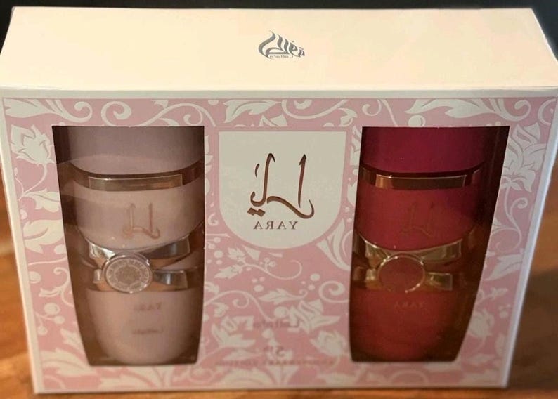 Peut inclure: Coffret cadeau avec deux contenants cylindriques. L'un est rose clair, l'autre rouge. Les deux ont des accents dor&eacute;s et une bande d&eacute;corative. La bo&icirc;te est blanche avec des motifs floraux roses et le mot "Yara" en &eacute;criture arabe.
