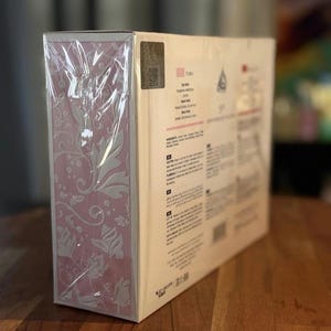 Peut inclure: Bo&icirc;te rectangulaire emball&eacute;e dans du plastique transparent. Un c&ocirc;t&eacute; pr&eacute;sente un motif floral rose et l'autre du texte, dont le nom "TARA". Un autocollant holographique est visible. Sur une surface en bois.