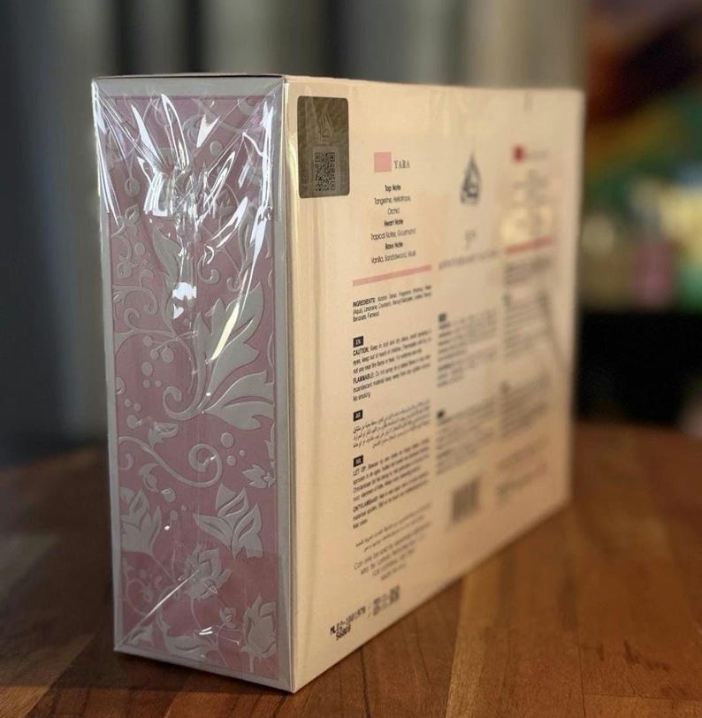 Peut inclure: Bo&icirc;te rectangulaire emball&eacute;e dans du plastique transparent. Un c&ocirc;t&eacute; pr&eacute;sente un motif floral rose et l'autre du texte, dont le nom "TARA". Un autocollant holographique est visible. Sur une surface en bois.