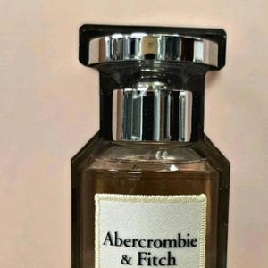 Może przedstawiać: Prostokątny flakon perfum ze srebrną nakrętką i ciemnobrązowym płynem w środku. Butelka ma białą etykietę z napisem "Abercrombie & Fitch AUTHENTIC". Tło jest w kolorze jasnor&oacute;żowym.