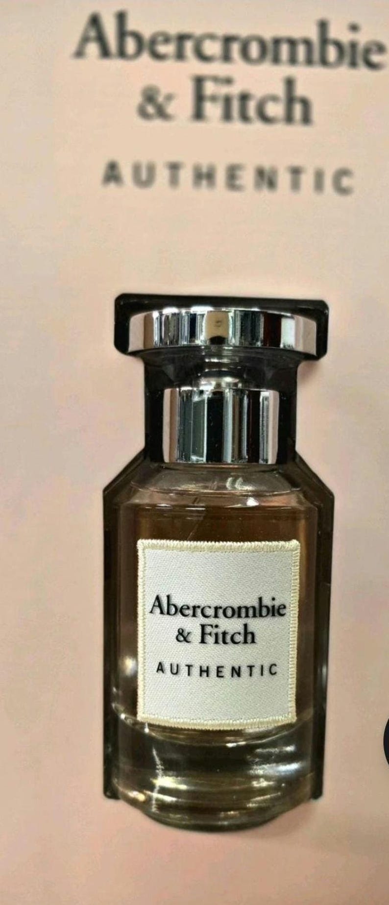 Może przedstawiać: Prostokątny flakon perfum ze srebrną nakrętką i ciemnobrązowym płynem w środku. Butelka ma białą etykietę z napisem "Abercrombie & Fitch AUTHENTIC". Tło jest w kolorze jasnor&oacute;żowym.