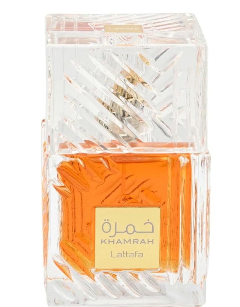 May include: A rectangular glass perfume bottle with a textured, faceted surface containing amber-colored liquid. A square gold label on the front features the text "KHAMRAH Lattafa" in English and Arabic script. The bottle has a clear, matching glass cap.
