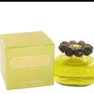 Puede incluir: Una caja amarilla y dorada con el texto "Covet by Marc Jacobs" en la parte delantera. La caja contiene un frasco de perfume de vidrio transparente con un tapón en forma de flor marrón. El frasco está lleno de un líquido amarillo. El frasco es de 100 ml.