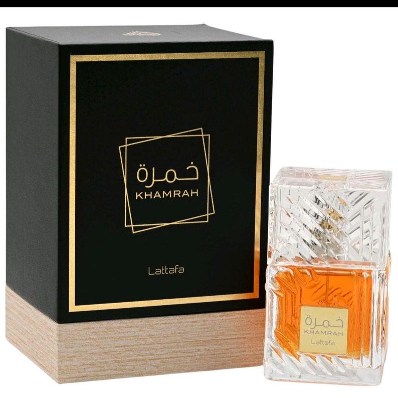 May include: A rectangular black and light wood-toned perfume box with gold trim and the text Khamrah Lattafa, accompanied by a clear, faceted glass perfume bottle containing amber liquid with a matching gold label featuring the same text.