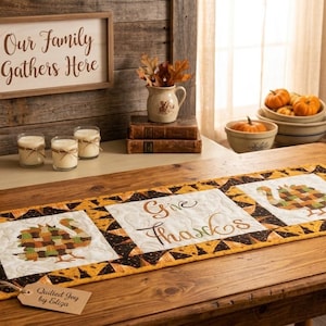 May include: A rectangular quilted table runner featuring a central white panel with the text Give Thanks in brown script, flanked by two white panels with patchwork turkey designs in orange, brown, and green, all bordered by a black and orange pattern.