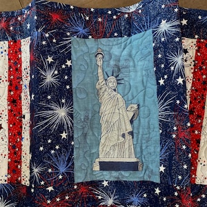 Puede incluir: Un edredón patriótico con la Estatua de la Libertad sobre un fondo azul claro. El edredón tiene un borde azul marino con estrellas blancas y fuegos artificiales rojos y blancos. El diseño del edredón recuerda a la bandera estadounidense.
