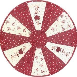 May include: A round, quilted placemat with a radial design. Alternating sections of burgundy fabric with white hearts and cream fabric with floral patterns. The word "LOVE" is embroidered in pink with a heart in the center of the word.