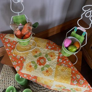 Op de afbeelding: Een Paasdecoratie met een bloemrijke, gewatteerde tafelloper in oranje, geel en wit. Twee metalen konijnenfiguren houden groene emmers met paaseieren en worteldecoraties vast. Groene kaarsen en een geweven placemat maken de scène compleet.