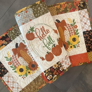 May include: Two quilted table runners featuring a patchwork border in autumn colors of orange, brown, and yellow. The center panels display embroidered pumpkins, sunflowers, and the text Hello Fall in cursive script, accented with decorative stitching.