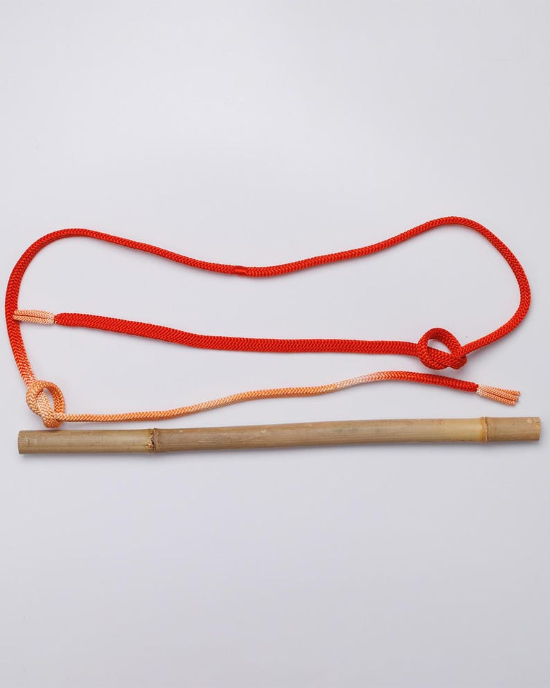 May include: A red and orange rope tied to a wooden stick. The rope is tied in a knot and the ends of the rope are frayed.