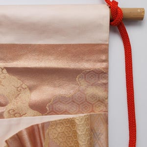 May include: A traditional Japanese hanging scroll featuring a pink and gold fabric design. The scroll is attached to a wooden dowel with a red cord.