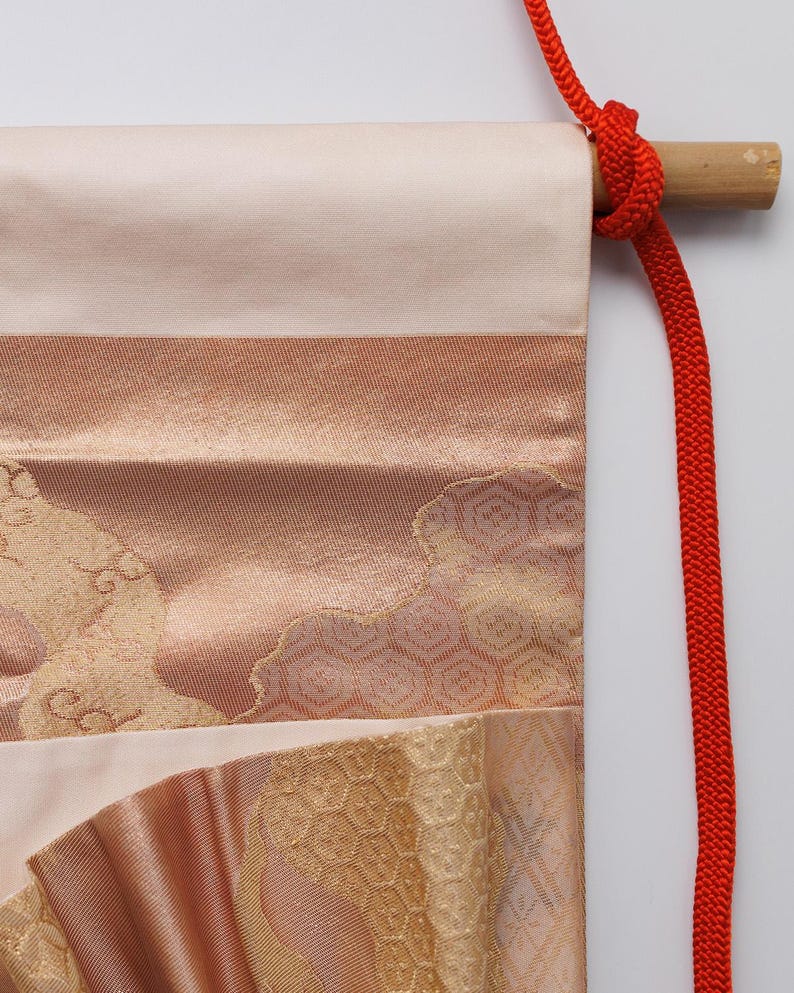 May include: A traditional Japanese hanging scroll featuring a pink and gold fabric design. The scroll is attached to a wooden dowel with a red cord.