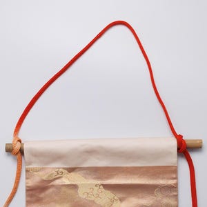 May include: A cream-coloured fabric banner with gold and orange patterns hangs from a wooden dowel with a red cord. The cord is tied in a knot at the top and hangs down on either side of the banner.