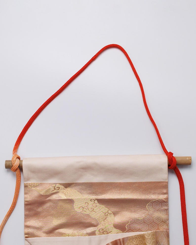 May include: A cream-coloured fabric banner with gold and orange patterns hangs from a wooden dowel with a red cord. The cord is tied in a knot at the top and hangs down on either side of the banner.