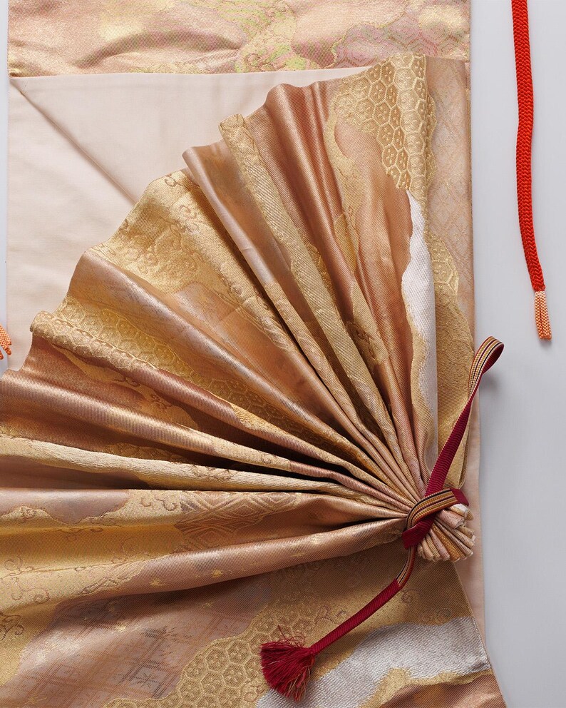 May include: A gold and pink fabric fan with a red tassel. The fan is folded and tied with a red ribbon.