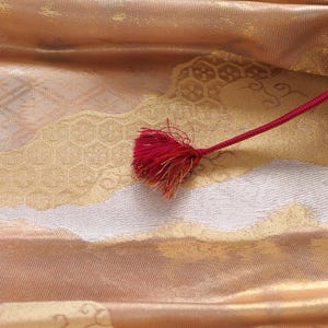 May include: A close-up of a gold and white fabric with a red tassel. The fabric has a woven pattern and a subtle sheen.
