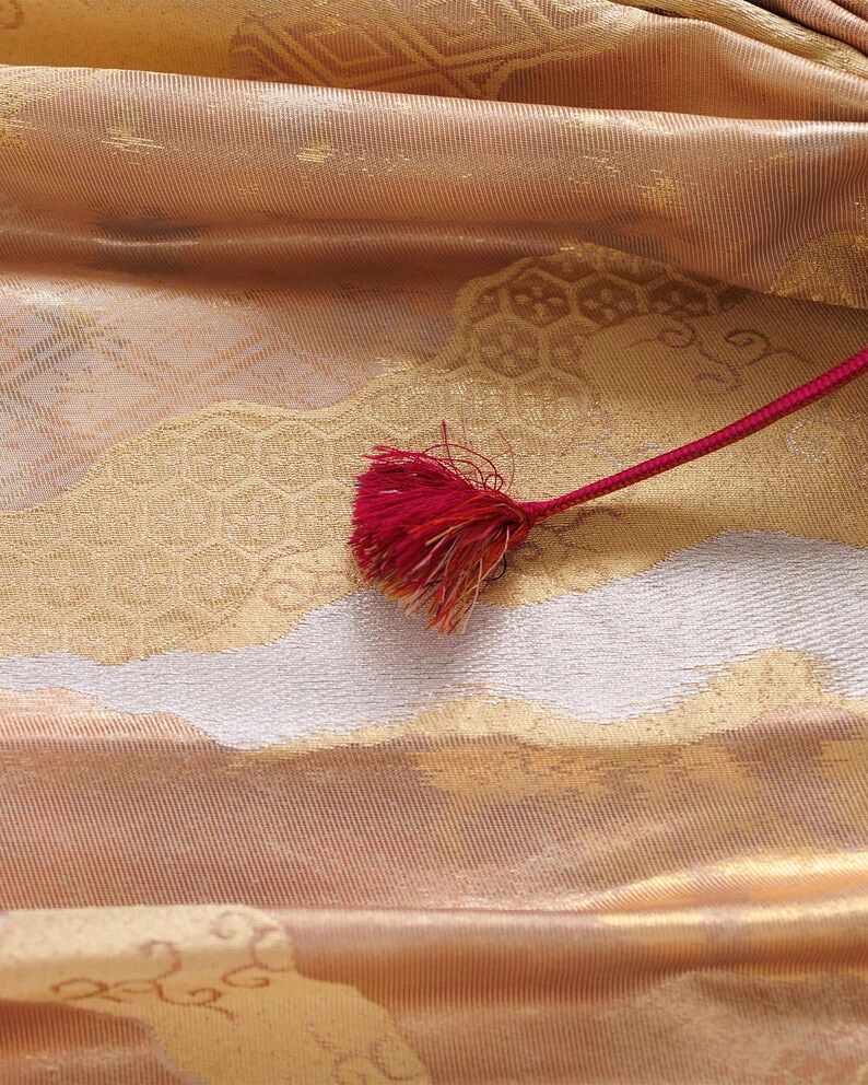 May include: A close-up of a gold and white fabric with a red tassel. The fabric has a woven pattern and a subtle sheen.