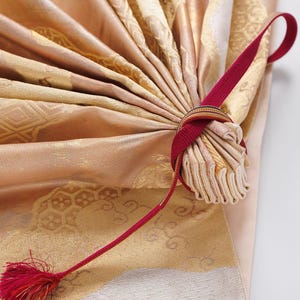 May include: A close-up of a gold and white fabric fan with a red ribbon tied around the handle. The fan is folded and the ribbon is tied in a bow.