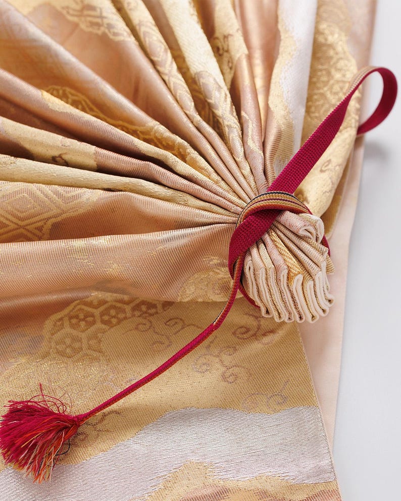 May include: A close-up of a gold and white fabric fan with a red ribbon tied around the handle. The fan is folded and the ribbon is tied in a bow.