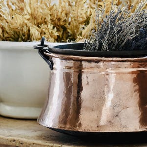 May include: A polished copper pot with a wide rim and dark metal handles, filled with dried purple lavender sprigs. The pot features a hammered texture and sits in front of a large white ceramic bowl containing dried beige foliage and folded linens.